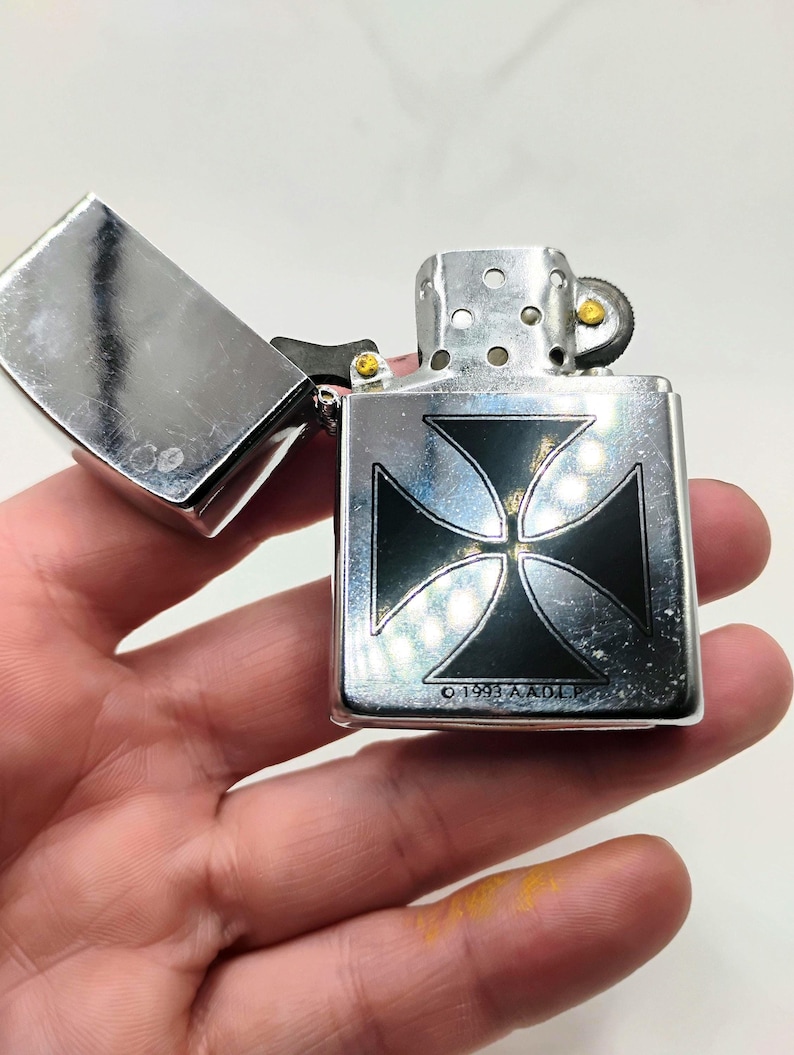 May include: A silver metal lighter featuring a black cross design. The lid is open, showing the inside. The text "&copy; 1993 A.A.O.L.P." is printed on the lighter.