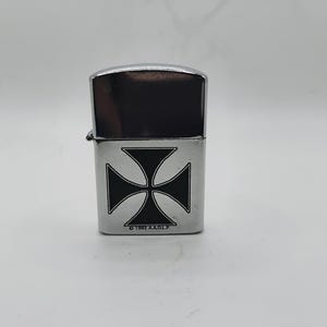 May include: A silver metal lighter featuring a black Maltese cross design. The top is a shiny, dark silver. The text "&copy; 1991 A.A.O.F." is printed at the bottom. The lighter is a classic design, suitable for everyday use.