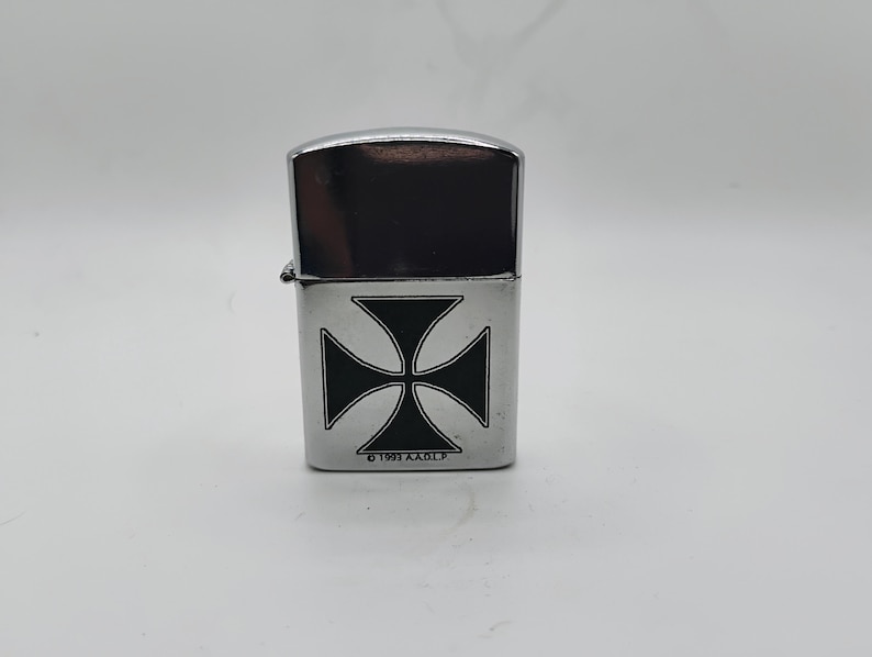 May include: A silver metal lighter featuring a black Maltese cross design. The top is a shiny, dark silver. The text "&copy; 1991 A.A.O.F." is printed at the bottom. The lighter is a classic design, suitable for everyday use.