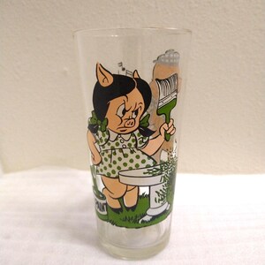 Op de afbeelding: Een helder glas met een cartoonafbeelding van een varkensmeisje dat een verfkwast vasthoudt en voor een verfpot staat. Het varkensmeisje draagt een groene stippenjurk en heeft zwart haar. Het glas maakt deel uit van een set glazen met verschillende cartoonfiguren.
