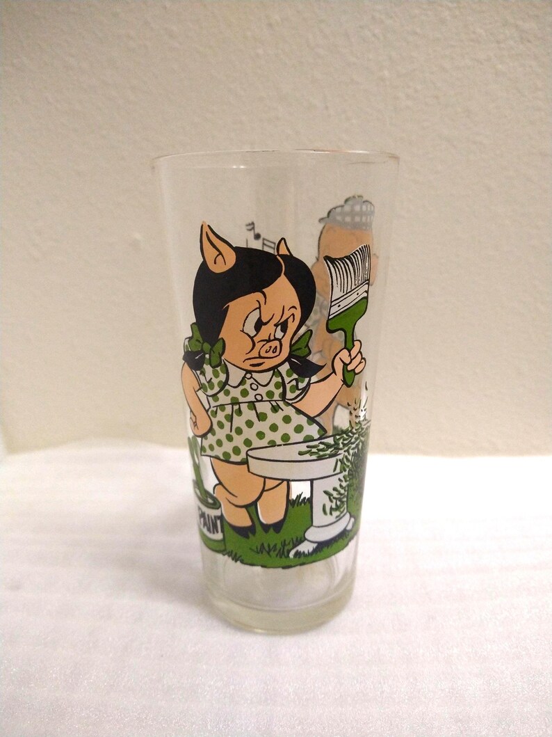 Op de afbeelding: Een helder glas met een cartoonafbeelding van een varkensmeisje dat een verfkwast vasthoudt en voor een verfpot staat. Het varkensmeisje draagt een groene stippenjurk en heeft zwart haar. Het glas maakt deel uit van een set glazen met verschillende cartoonfiguren.