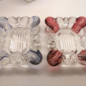 May include: Two glass ashtrays with a decorative design. One ashtray is clear with a blue tint, and the other is clear with a red tint.