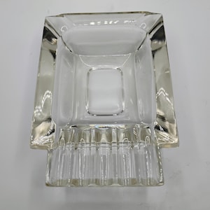 May include: A clear, rectangular glass ashtray with a square interior. The ashtray has a ribbed base and a thick, beveled rim. The design is simple and elegant, suitable for a modern or vintage aesthetic.