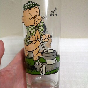 Op de afbeelding: Een helder glas met een cartoon afbeelding van Porky Pig die de grasmat maait. Hij draagt een groen shirt met witte stippen, een groene strik en een geruite hoed. Hij blaast een wolk lucht uit met muzieknootjes die uit zijn mond komen.
