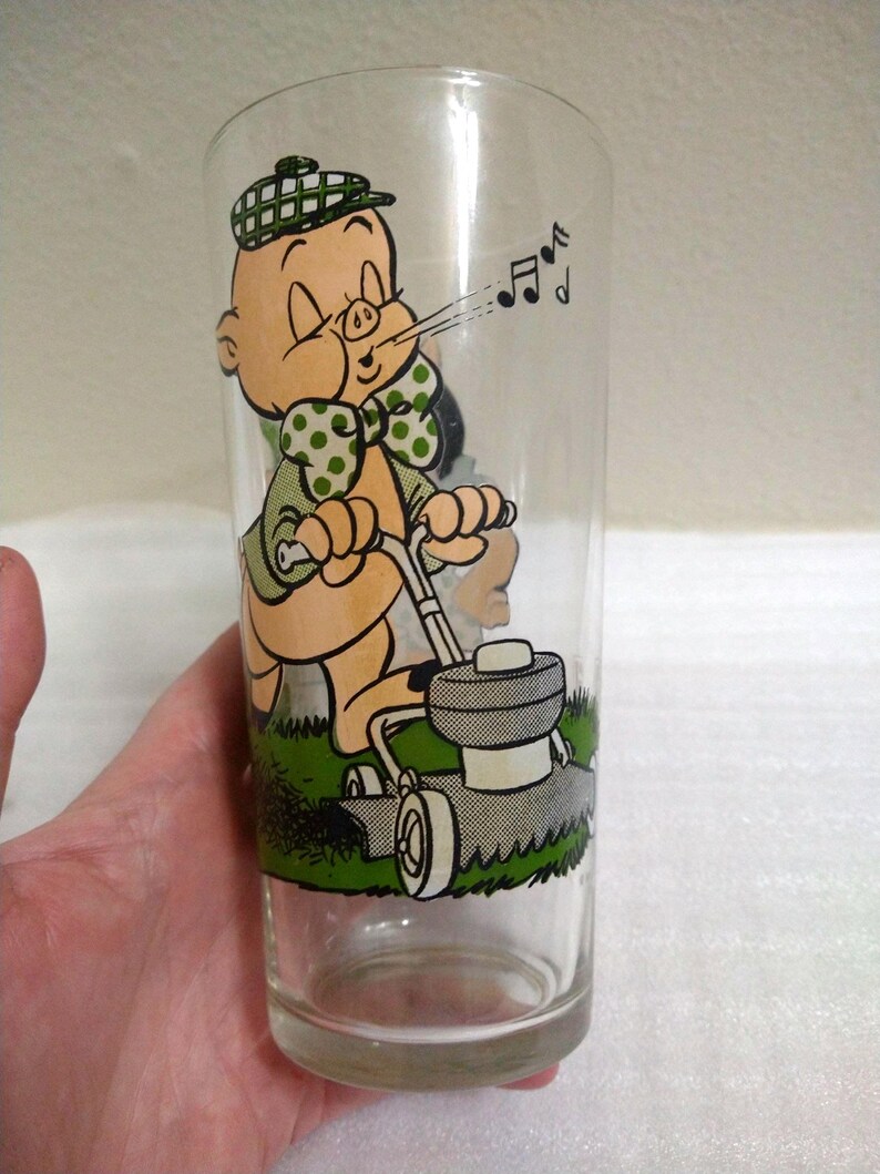 Op de afbeelding: Een helder glas met een cartoon afbeelding van Porky Pig die de grasmat maait. Hij draagt een groen shirt met witte stippen, een groene strik en een geruite hoed. Hij blaast een wolk lucht uit met muzieknootjes die uit zijn mond komen.