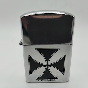 May include: A chrome lighter featuring a black Maltese cross emblem. The lighter has a polished, reflective finish. The text "&copy; 1993 A.A.D.L.P." is printed at the base of the lighter.