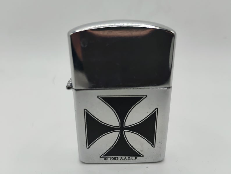 May include: A chrome lighter featuring a black Maltese cross emblem. The lighter has a polished, reflective finish. The text "&copy; 1993 A.A.D.L.P." is printed at the base of the lighter.