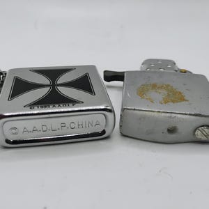 May include: Close-up of three silver lighters. One lighter features a black and white cross design. The bottom of the lighter reads "A.A.D.L.P. CHINA". The other lighters are plain silver.