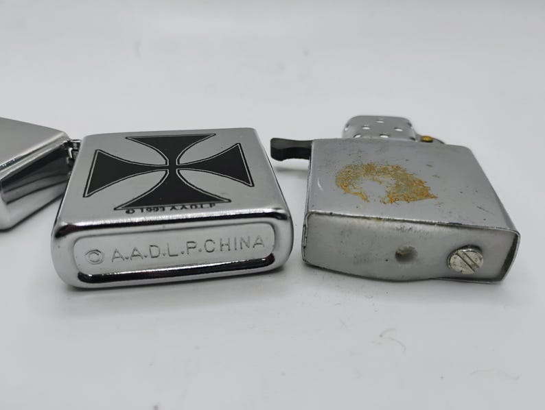 May include: Close-up of three silver lighters. One lighter features a black and white cross design. The bottom of the lighter reads "A.A.D.L.P. CHINA". The other lighters are plain silver.