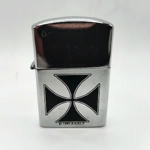 May include: A silver metal lighter featuring a black Maltese cross design. The lighter has a shiny, reflective top and the text "&copy; 1993 A.A.D.L.P." at the bottom. A classic accessory.