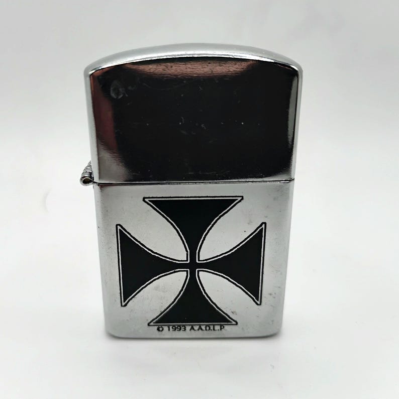 May include: A silver metal lighter featuring a black Maltese cross design. The lighter has a shiny, reflective top and the text "&copy; 1993 A.A.D.L.P." at the bottom. A classic accessory.