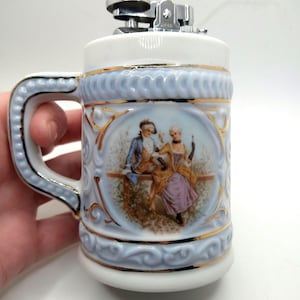 May include: A white ceramic lighter shaped like a beer stein with a blue and gold design. The stein features a couple in a romantic scene. The lighter has a silver top with a working flint.