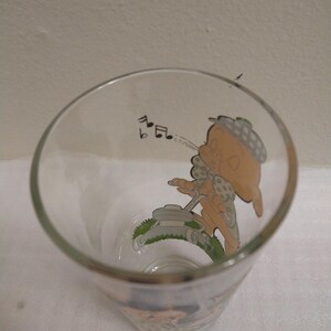 Op de afbeelding: Een helder glas met een cartoonafbeelding van een personage dat de grasmat maait terwijl het een melodie fluit. Het personage draagt een hoed en heeft een gestippeld patroon op zijn kleding. Het glas is leeg.