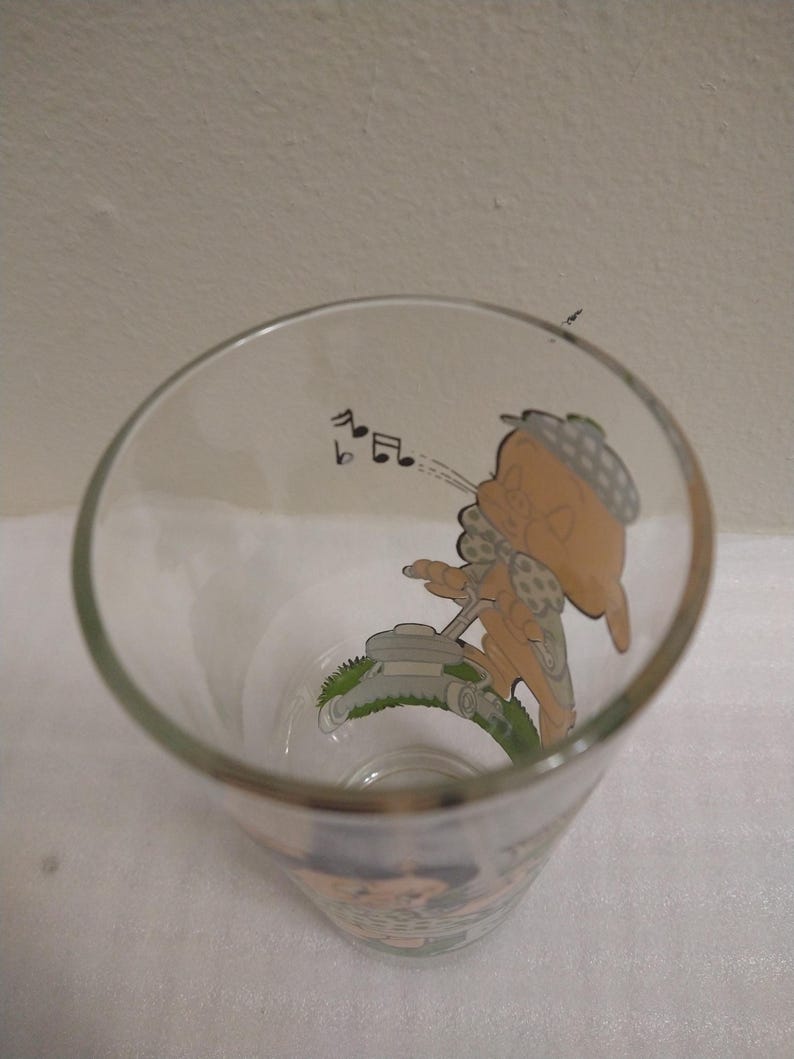 Op de afbeelding: Een helder glas met een cartoonafbeelding van een personage dat de grasmat maait terwijl het een melodie fluit. Het personage draagt een hoed en heeft een gestippeld patroon op zijn kleding. Het glas is leeg.