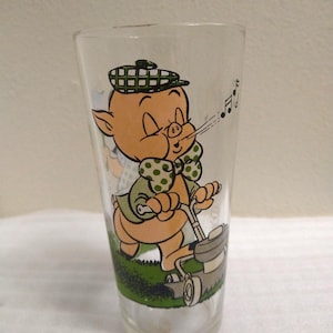 Op de afbeelding: Een glas met een cartoon afbeelding van een varken dat een hoed draagt en muzieknoten blaast terwijl het op een grasmaaier rijdt. Het varken draagt een groen shirt met witte stippen en een groen-wit geruite hoed.