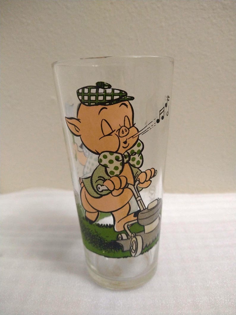 Op de afbeelding: Een glas met een cartoon afbeelding van een varken dat een hoed draagt en muzieknoten blaast terwijl het op een grasmaaier rijdt. Het varken draagt een groen shirt met witte stippen en een groen-wit geruite hoed.