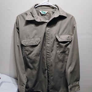May include: A tan button-down shirt with two chest pockets and long sleeves, hanging on a white hanger. The shirt has a collar and a label at the neck. The fabric appears to be a durable, workwear style.