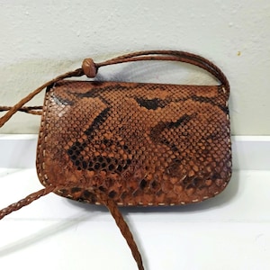 May include: A small, brown leather crossbody bag with a snakeskin pattern. The bag has a braided strap and a small, rounded flap closure. The stitching is visible around the edges of the bag. The bag is approximately 6 inches wide and 4 inches tall.