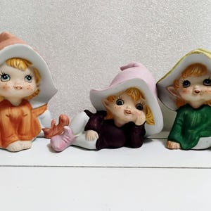 May include: Three ceramic elf figurines with large hats and smiling faces. One elf wears an orange outfit, another a dark purple outfit, and the third a green outfit. Each figurine has a unique pose.