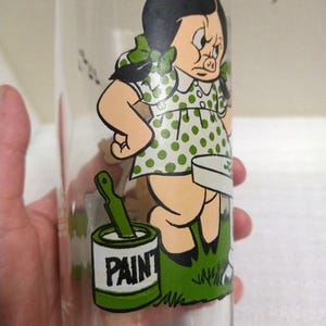 Op de afbeelding: Een vintage glas met een cartoon afbeelding van een varkensmeisje in een groene stippenjurk die een kwast en een blik verf vasthoudt waarop "PAINT" staat.