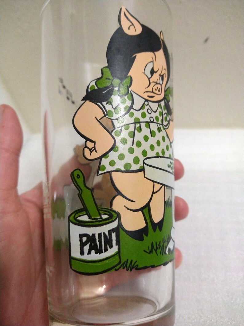 Op de afbeelding: Een vintage glas met een cartoon afbeelding van een varkensmeisje in een groene stippenjurk die een kwast en een blik verf vasthoudt waarop "PAINT" staat.