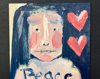 May include: A square wooden panel featuring an acrylic painting of a face with dark hair, pink cheeks, and two red hearts on the right side. The word Peace is painted in dark blue letters at the bottom against a light background with dark blue edges.