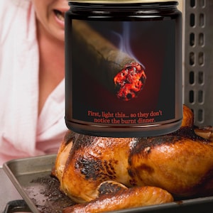 May include: A glass jar candle with a label that reads "First, light this... so they don't notice the burnt dinner." The label features a close-up image of a lit cigar with smoke rising from it. The candle is sitting on top of a burnt turkey in a baking pan.