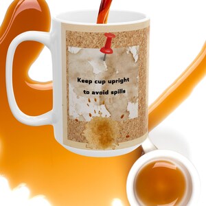 Op de afbeelding: Witte keramische koffiemok met een kurkachtig ontwerp met een rode duimspijker en een bruine vlek. De tekst op de mok luidt "Keep cup upright to avoid spills".