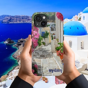 以下が含まれることがあります： ギリシャの村の美しい景色が描かれたスマートフォンケース。白い建物、青いドーム、ピンクの花が咲く石畳の道が特徴です。「αγάπη」という言葉がケースに印刷されています。