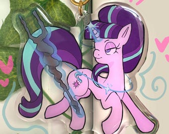 May include: A clear acrylic keychain featuring a cartoon pony with a light pink body, purple and teal mane, and a horn. The pony holds a dark, twisted staff and has a star-shaped mark on its flank. It includes a gold-colored star-shaped clasp.