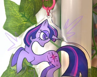 May include: A clear acrylic keychain featuring a purple pony character with a dark blue and purple mane and tail. The pony has a star-shaped cutie mark on its flank and a small horn. It is attached to a bright pink plastic swivel clasp hook.