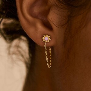 May include: Gold-colored stud earring with a starburst design and a small, light pink center stone. A delicate gold chain hangs from the earring. The text "PEARL ECLIPSE DESIGNS" is at the bottom of the image.