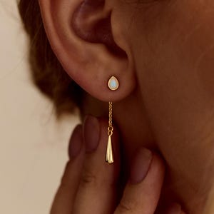 Peut inclure: Boucles d'oreilles dorées avec un design en forme de goutte d'eau. L'une est une petite puce avec une pierre blanche, l'autre pend avec une chaîne et un charme conique. Les boucles d'oreilles sont portées sur une oreille.