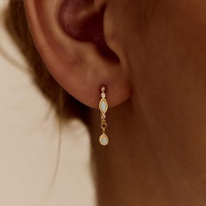 May include: Close-up of a gold earring with a delicate design. The earring features a small, round stud with a dangling chain holding two oval-shaped, white gemstones. The text "PEARL ECLIPSE DESIGNS" is at the bottom.