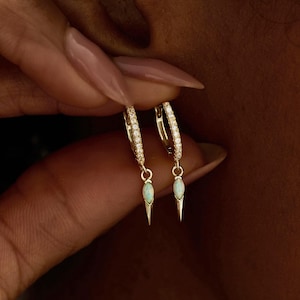 Könnte beinhalten: Goldene Creolen mit kleinen weißen Edelsteinen und hängenden, hellgrünen, spitzförmigen Anhängern. Die Ohrringe werden ans Ohr gehalten. Der Text "PEARL ECLIPSE DESIGNS" befindet sich unten im Bild.