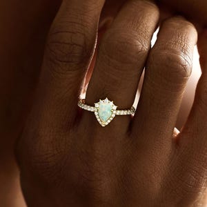 May include: A gold ring featuring a pear-shaped, pale gemstone, encircled by smaller, clear stones. The ring adorns a finger, with the hand and part of the face in view. The ring is set against a soft backdrop.