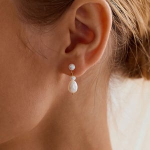 May include: A gold earring featuring a small round white pearl stud connected to a dangling teardrop-shaped white pearl. The earring is worn on the earlobe. The text PEARL ECLIPSE DESIGNS is printed in white at the bottom of the image.