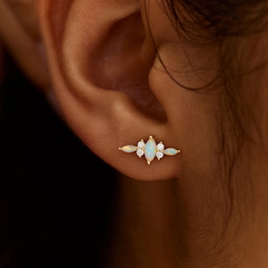 May include: A gold-colored earring with an opal center stone and small white gemstones. The earring is a bar style with a symmetrical design. The text "PEARL ECLIPSE DESIGNS" is at the bottom of the image.