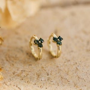 May include: Two gold-coloured hoop earrings, each featuring three dark teal gemstones. The earrings are set against a neutral, textured backdrop, with soft, blurred elements in the background.