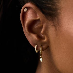 May include: Close-up of an ear with gold earrings. A small stud earring is in the upper ear, and two gold hoop earrings are in the lower ear. One hoop has small crystals, and the other has a white teardrop pendant. The text "PEARL ECLIPSE DESIGNS" is at the bottom.