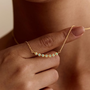 Peut inclure: Un collier en or délicat orné d'une série de petites opales rondes. Les opales sont disposées en ligne incurvée. Le collier est présenté par une main. Le nom de la marque "PEARL ECLIPSE DESIGNS" est en bas.