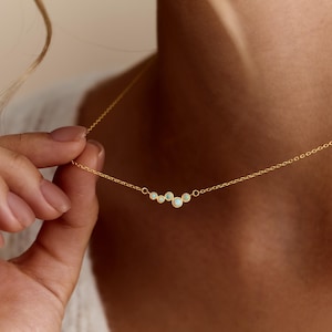 May include: A delicate gold chain necklace featuring a cluster of five small, round, iridescent opal-like stones set in gold bezels. The stones are arranged in a slightly curved, asymmetrical line, suspended from a thin, polished gold link chain.