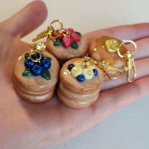 May include: Miniature pancake keychains with various toppings, including blueberries, strawberries, and bananas. Each keychain features a golden clasp. The pancakes are stacked and appear to be made of clay or a similar material.