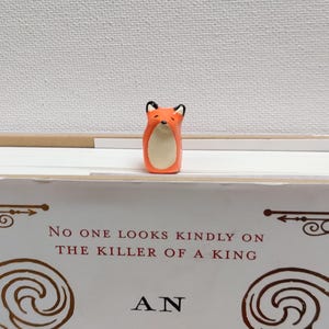 May include: A small orange fox figurine with white accents, sitting on a book with the text "No one looks kindly on the killer of a king. An..."