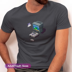 May include: A dark gray t-shirt with a pixelated illustration of a Super Nintendo Entertainment System (SNES) console, a controller, and a television screen displaying a video game. The text "1990" is visible on the t-shirt.