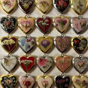 Puede incluir: Una colección de colgantes en forma de corazón con flores secas encapsuladas en resina. Los colgantes tienen marcos de metal dorado, plateado y negro. Cada colgante presenta una disposición única de flores secas coloridas.