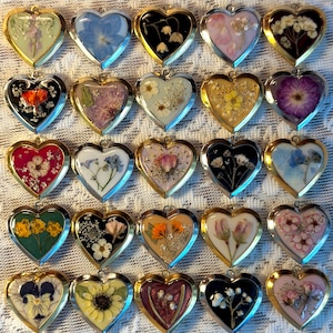 May include: A collection of twenty-five heart-shaped pendants arranged in a grid. Each pendant features a unique pressed flower or botanical element encased in clear resin with a gold-toned metal frame and a small loop at the top for hanging.