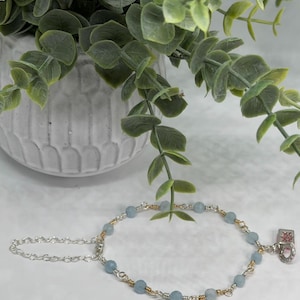 May include: A delicate bracelet featuring light blue beads strung on silver and gold-toned wire. A small charm with a red and white design hangs from the bracelet. The bracelet is displayed in front of a white pot with green foliage.