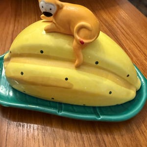 May include: A ceramic butter dish featuring a yellow starfruit shape with black dots, topped with an orange monkey figurine. The dish sits on a green leaf-shaped base, creating a whimsical and decorative kitchen item.