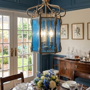 May include: A hanging lantern pendant light featuring a weathered metallic frame with ornate leaf details and six panels of textured blue glass. The fixture is suspended by a metal chain and houses multiple internal light bulbs.